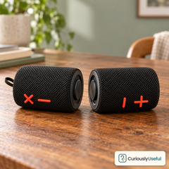 Two black magnetic Bluetooth speaker sections lying on a timber table with their joining ends facing each other and a visible Curiously Useful logo.